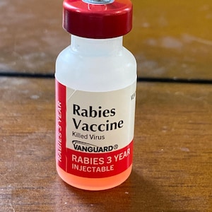 May include: A clear glass vial with a red cap and a silver loop at the top. The vial is labeled "Rabies Vaccine" with the text "Vanguard" and "Rabies 3 Year Injectable".