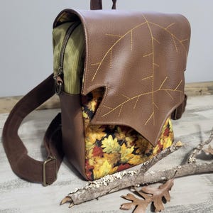 May include: A brown leather backpack with a large leaf-shaped flap. The flap has a stitched design of veins and is accented with a gold zipper. The backpack has a brown fabric lining with a pattern of autumn leaves in shades of yellow, orange, and red.