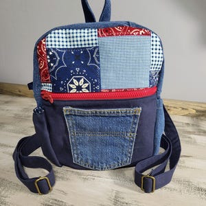May include: A small blue denim backpack with a patchwork design on the front flap. The patchwork includes red, white, and blue bandana print fabric, gingham fabric, and denim fabric. The backpack has a zippered pocket on the front and a large denim pocket on the bottom.