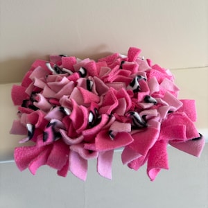 Könnte beinhalten: Eine handgefertigte Haustiermatte in Rosatönen mit Akzenten in Schwarz und Weiß. Die Matte besteht aus weichem Fleece-Stoff, wobei Stoffstreifen miteinander verknotet sind, um eine strukturierte Oberfläche zu schaffen. Die Matte ist für kleine Haustiere konzipiert.