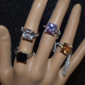 May include: Four silver rings with square-shaped gemstones. The rings feature a clear, black, light purple, and orange gemstone.
