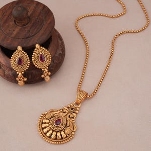 May include: A gold-toned jewelry set featuring a pendant necklace and matching earrings. The pendant and earrings have a teardrop shape with a central red gemstone and intricate, ornate detailing. The necklace chain is a delicate, textured design.