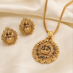 May include: A gold-toned jewelry set consisting of a pendant necklace and matching stud earrings. Each piece features an intricate embossed figure seated on a lotus flower, surrounded by ornate filigree detailing and a textured border finish.