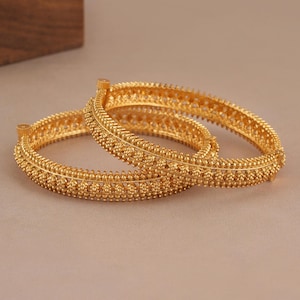 May include: Two circular gold bangles featuring a textured surface with intricate beaded detailing and a small cylindrical clasp mechanism on the side of each piece. The bangles have a polished metallic finish and a consistent round shape.