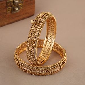 May include: Two gold-toned bangles featuring a textured surface with repeating bead-like patterns and a central lattice design. Each circular bracelet includes a small hinged clasp mechanism for opening and closing, showcasing a metallic finish.