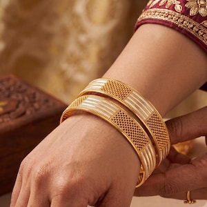 May include: Two gold bangles featuring a repeating pattern of textured, dotted gold sections alternating with smooth, polished silver-toned vertical stripes. The bracelets have a rounded profile and a high-shine finish, creating a contrast between the yellow gold and silver-toned elements.