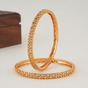 May include: Two gold-toned bangles featuring a textured, repeating pattern along the outer surface. The bangles are circular in shape with a metallic finish, one positioned vertically and the other resting horizontally on a flat surface.