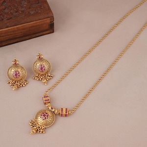 May include: A gold-toned jewelry set featuring a necklace and matching earrings. The necklace has a pendant with a central red stone surrounded by intricate detailing, and a chain. The earrings mirror the pendant design. The set is displayed on a neutral background.