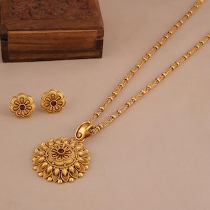 May include: A gold-colored jewelry set featuring a pendant necklace and matching stud earrings. The pendant and earrings have a floral design with a small, red gemstone in the center. The necklace chain is a delicate, beaded style.