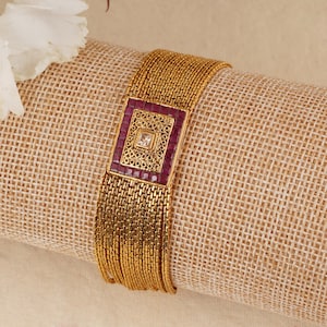 May include: A gold bracelet with a square-shaped centerpiece set with red and white stones.