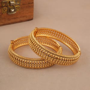 May include: Two gold-colored bangles with intricate beaded and openwork designs. The bangles are displayed on a neutral surface, showcasing their detailed craftsmanship. A wooden box is partially visible in the background.