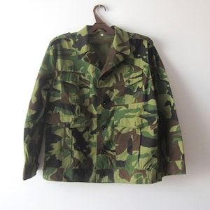 May include: A green, brown, and black camouflage jacket with a button closure and multiple pockets. The jacket is hanging on a wooden hanger against a white background.