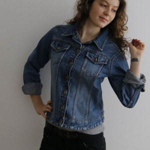 Könnte beinhalten: Eine Frau trägt eine blaue Jeansjacke mit Knöpfen und ein hellgraues Hemd darunter. Sie trägt eine schwarze Jeans und hat ihre Haare nach hinten gebunden.