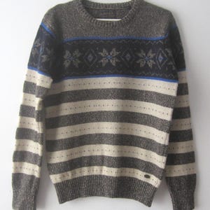 Puede incluir: Un suéter gris y blanco a rayas con un patrón de copo de nieve azul y negro alrededor del cuello. El suéter tiene cuello redondo y mangas largas.