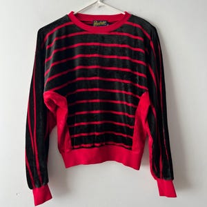 Può includere: Felpa a righe nere e rosse con maniche lunghe. La felpa ha girocollo, polsini e orlo inferiore rossi. Il marchio "Panther" è visibile sull'etichetta. Il tessuto sembra essere un materiale morbido e vellutato.