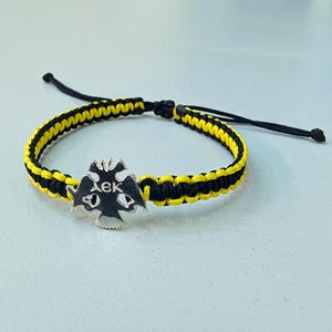 Op de afbeelding: Een zwart en geel gevlochten armband met een zilveren bedel. De bedel heeft een dubbelkoppige adelaar met Griekse letters. De armband is verstelbaar met zwarte koorden.