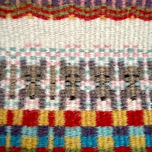 May include: Close-up of a woven textile featuring a vibrant mix of colours and patterns. The design incorporates horizontal bands of red, purple, blue, and yellow, with a repeating bear-like motif in the centre. The texture appears soft and slightly fuzzy.