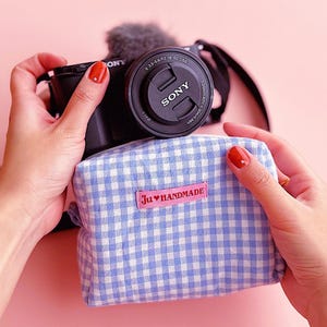 May include: A light blue and white gingham fabric pouch featuring a rectangular pink label with the text Ju♥HANDMADE in red. The pouch is positioned in front of a black Sony camera with a lens cap attached, showing technical lens specifications.