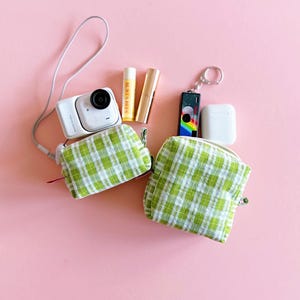 May include: A selection of small accessories on a pink backdrop. The image shows a white digital camera, two lip balms, a rainbow keychain, a white wireless earbud case, and two green and white plaid pouches.