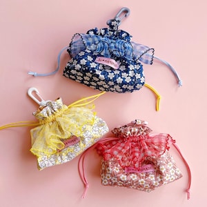 May include: Three small drawstring pouches in blue, yellow, and pink, each with a floral pattern and a ruffled trim. The blue pouch has a blue and white floral pattern, the yellow pouch has a yellow trim, and the pink pouch has a red trim. Each pouch has a tag that says "Ju Handmade".