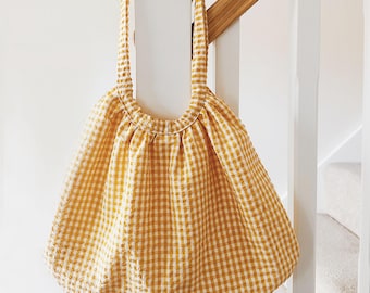 May include: A soft, unstructured tote bag made from a yellow and white gingham checkered fabric. The bag features a single integrated handle that forms a circular opening at the top, with gathered fabric creating a rounded, voluminous silhouette.