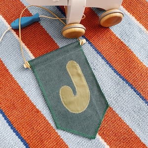 Puede incluir: Un juguete de hipopótamo de madera blanca para tirar, con ruedas de madera y un pez azul sujeto con una cuerda. Una bandera verde con una "J" beige está en primer plano. Los objetos están sobre una alfombra a rayas naranja, azul y blanca.