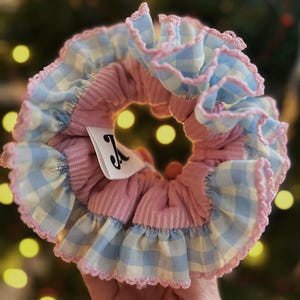 May include: A handmade scrunchie with a pink ribbed centre and blue and white gingham ruffle trim. The scrunchie has a small white tag with a black initial. The background is blurred with bokeh lights.