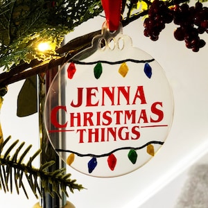 May include: A clear, round Christmas bauble with the words "JENNA CHRISTMAS THINGS" in red. It features a string of colourful lights and hangs from a red ribbon. Festive decoration for the holiday season.