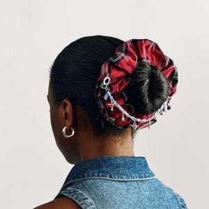 Puede incluir: Una goma para el pelo a cuadros rojos y negros con una cadena decorativa y abalorios. La goma para el pelo se lleva en un mo&ntilde;o. La persona lleva un chaleco vaquero y pendientes de aro dorados. El fondo es blanco liso.