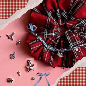Puede incluir: Una goma para el pelo a cuadros rojos con una cadena azul y abalorios, incluyendo corazones, coronas y llaves. Hay estrellas y coronas plateadas y una cinta azul esparcidas. El fondo es rosa con un patr&oacute;n de cuadros rojos y blancos.