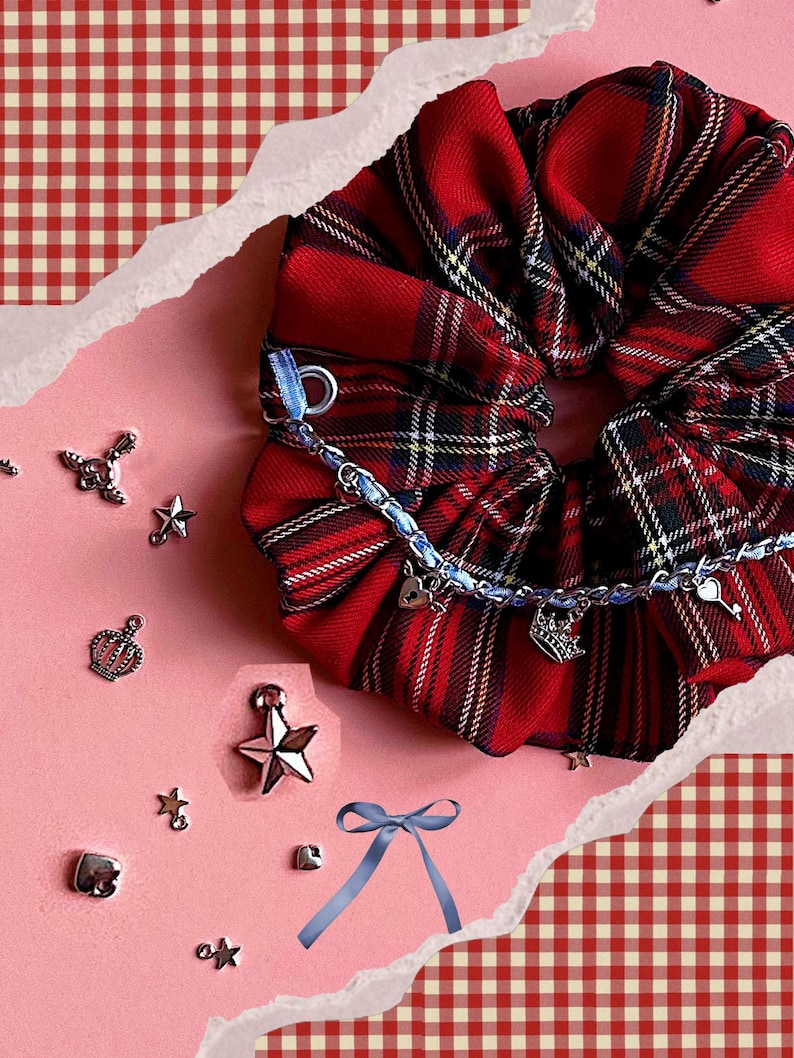 Puede incluir: Una goma para el pelo a cuadros rojos con una cadena azul y abalorios, incluyendo corazones, coronas y llaves. Hay estrellas y coronas plateadas y una cinta azul esparcidas. El fondo es rosa con un patr&oacute;n de cuadros rojos y blancos.