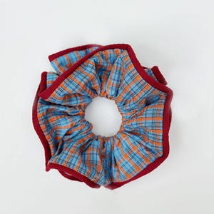 May include: A blue and orange plaid scrunchie with a red border. The hair accessory is circular with a hole in the center. The plaid pattern is made up of thin lines of blue and orange. The scrunchie is a hair accessory.