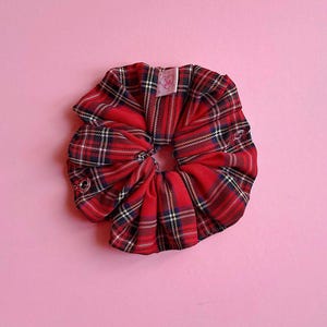 Puede incluir: Una goma para el pelo a cuadros rojos con detalles en negro, blanco y azul marino. La goma para el pelo es de tela y tiene una peque&ntilde;a etiqueta. El fondo es de color rosa liso.