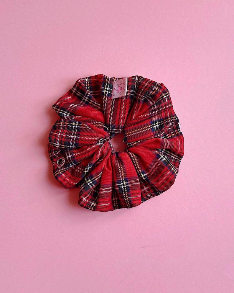 Puede incluir: Una goma para el pelo a cuadros rojos con detalles en negro, blanco y azul marino. La goma para el pelo es de tela y tiene una peque&ntilde;a etiqueta. El fondo es de color rosa liso.