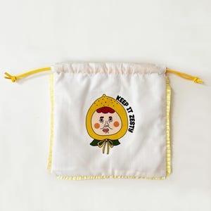 May include: White drawstring bag with a yellow ruffle trim and yellow drawstring. The bag features a cartoon illustration of a face wearing a yellow lemon-shaped hood with the text "KEEP IT ZESTY".