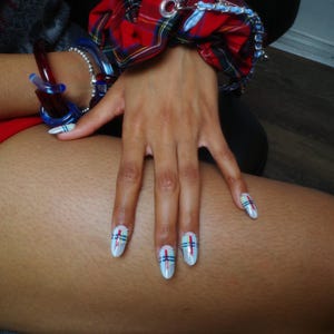 Puede incluir: Una mano con u&ntilde;as blancas, cada una adornada con un dise&ntilde;o de l&iacute;neas rojas, azules y negras. Tambi&eacute;n se ve una goma para el pelo a cuadros rojos y negros y pulseras azules. Las u&ntilde;as son el punto focal, mostrando un estilo de arte de u&ntilde;as &uacute;nico.