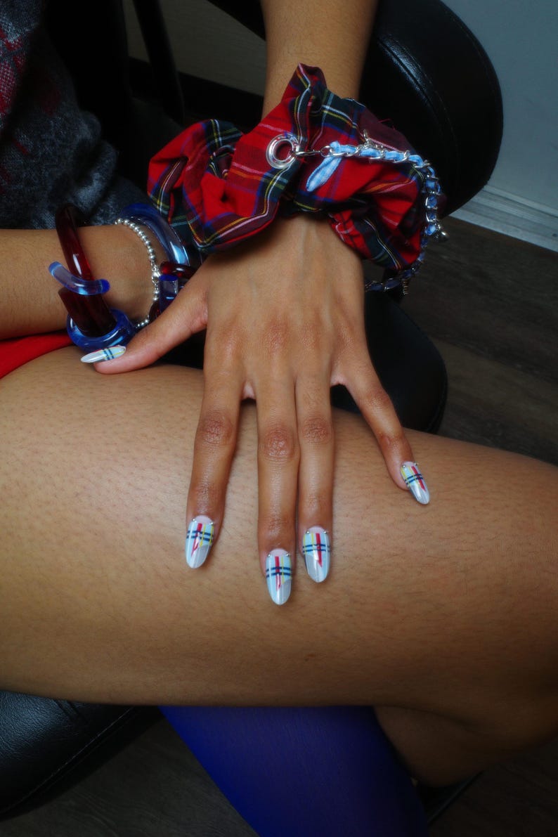 Puede incluir: Una mano con u&ntilde;as blancas, cada una adornada con un dise&ntilde;o de l&iacute;neas rojas, azules y negras. Tambi&eacute;n se ve una goma para el pelo a cuadros rojos y negros y pulseras azules. Las u&ntilde;as son el punto focal, mostrando un estilo de arte de u&ntilde;as &uacute;nico.