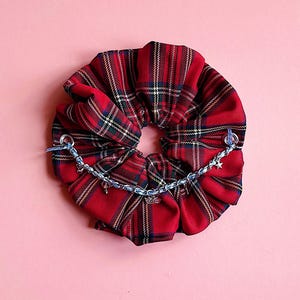 Puede incluir: Una goma para el pelo a cuadros rojos con una cadena azul y abalorios de estrella. La goma para el pelo est&aacute; hecha de una tela suave y tiene un dise&ntilde;o fruncido. La cadena est&aacute; unida a la goma para el pelo con peque&ntilde;os lazos azules. El fondo es rosa liso.
