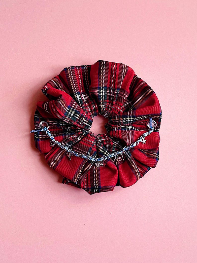 Puede incluir: Una goma para el pelo a cuadros rojos con una cadena azul y abalorios de estrella. La goma para el pelo est&aacute; hecha de una tela suave y tiene un dise&ntilde;o fruncido. La cadena est&aacute; unida a la goma para el pelo con peque&ntilde;os lazos azules. El fondo es rosa liso.
