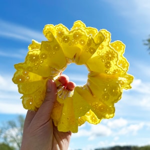 Peut inclure: Un chouchou en tissu jaune avec une délicate bordure en dentelle et un design plissé. Le chouchou est tenu contre un ciel bleu vif.