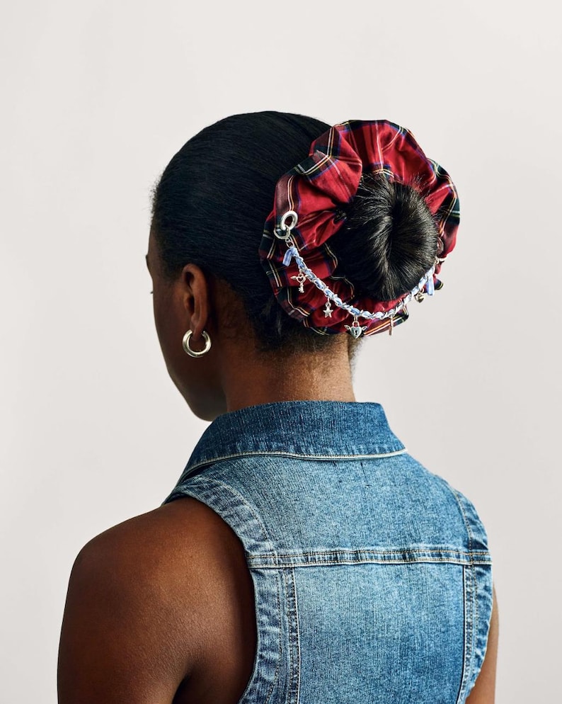 Puede incluir: Una goma para el pelo a cuadros rojos y negros con una cadena decorativa y abalorios. La goma para el pelo se lleva en un mo&ntilde;o. La persona lleva un chaleco vaquero y pendientes de aro dorados. El fondo es blanco liso.