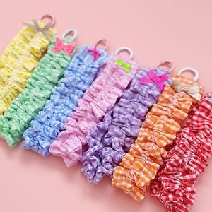 May include: A selection of fabric scrunchies in a variety of colours, including yellow, green, blue, pink, purple, orange, and red gingham. Each scrunchie has a matching bow and a white loop. The scrunchies are displayed on a pink background.