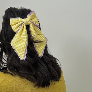 Può includere: Un fiocco di vichy giallo e bianco con un bordo viola. Il fiocco è legato in un grande nodo e viene indossato nei capelli di una persona.