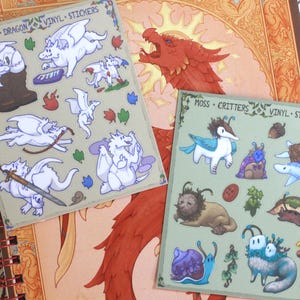 May include: Two sticker sheets featuring whimsical illustrations of dragons and fantasy creatures. One sheet is labeled "Little Dragon Vinyl Stickers" and the other "Moss Critters Vinyl Stickers". The stickers are on a light green background.