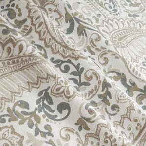 May include: Close-up of a fabric with a paisley pattern. The design features intricate floral and scroll motifs in shades of white, gray, and brown. The fabric has a slightly wrinkled texture, suggesting it is a soft material.