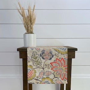 May include: A floral table runner with a beige background and a colorful floral pattern in shades of red, orange, blue, and green. The runner is on a wooden table with a vase of dried wheat.