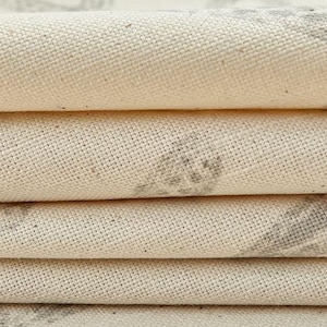 May include: A stack of folded off-white canvas fabric featuring a visible woven texture. The fabric displays faint, muted gray patterned markings across the surface, with the layers creating distinct horizontal folds that highlight the material.