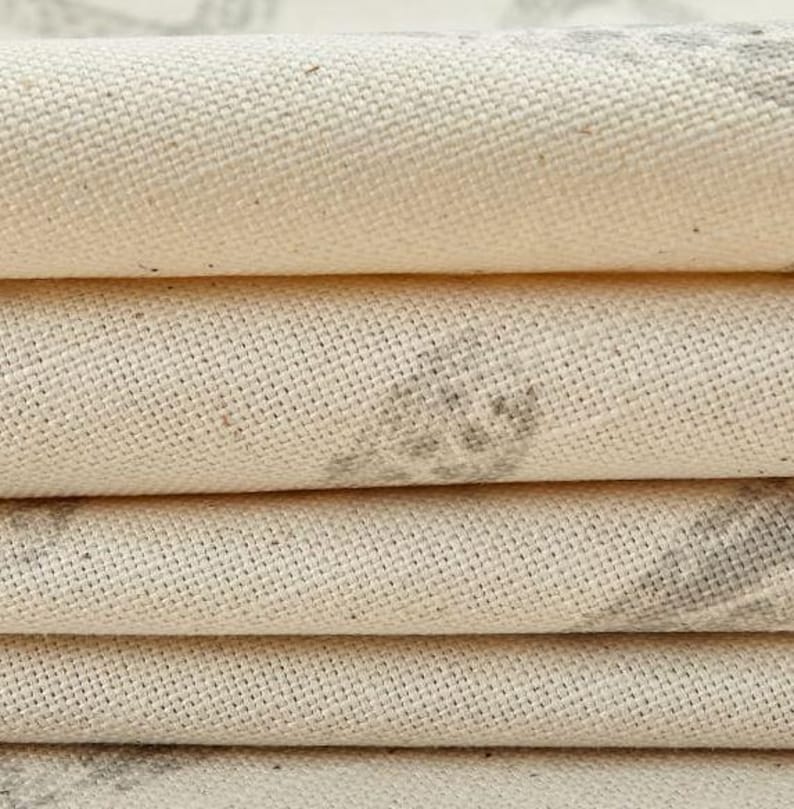 May include: A stack of folded off-white canvas fabric featuring a visible woven texture. The fabric displays faint, muted gray patterned markings across the surface, with the layers creating distinct horizontal folds that highlight the material.