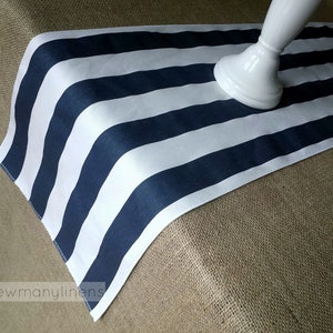 May include: A white and navy blue striped table runner. The runner is made of fabric and has a slightly textured surface. The runner is lying on a hessian surface.