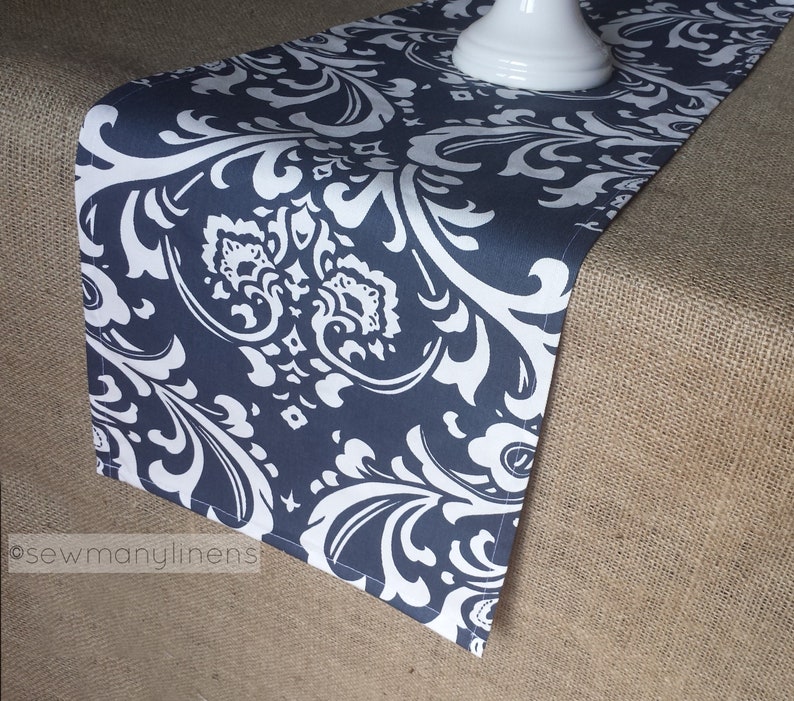 May include: A navy blue table runner with a white damask pattern. The runner is lying on a hessian surface.